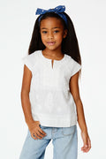 A young girl with long dark hair wears a blue headband, light blue jeans, and the Roller Rabbit Girls Faith Top in white with vibrant embroidery, standing against a plain white background and smiling slightly.