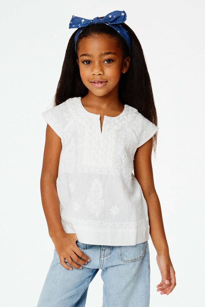 A young girl with long dark hair wears a blue headband, light blue jeans, and the Roller Rabbit Girls Faith Top in white with vibrant embroidery, standing against a plain white background and smiling slightly.