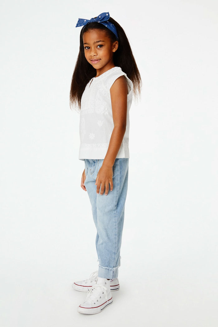 A young girl with long dark hair wears a blue headband, light blue jeans, and the Roller Rabbit Girls Faith Top in white with vibrant embroidery, standing against a plain white background and smiling slightly.