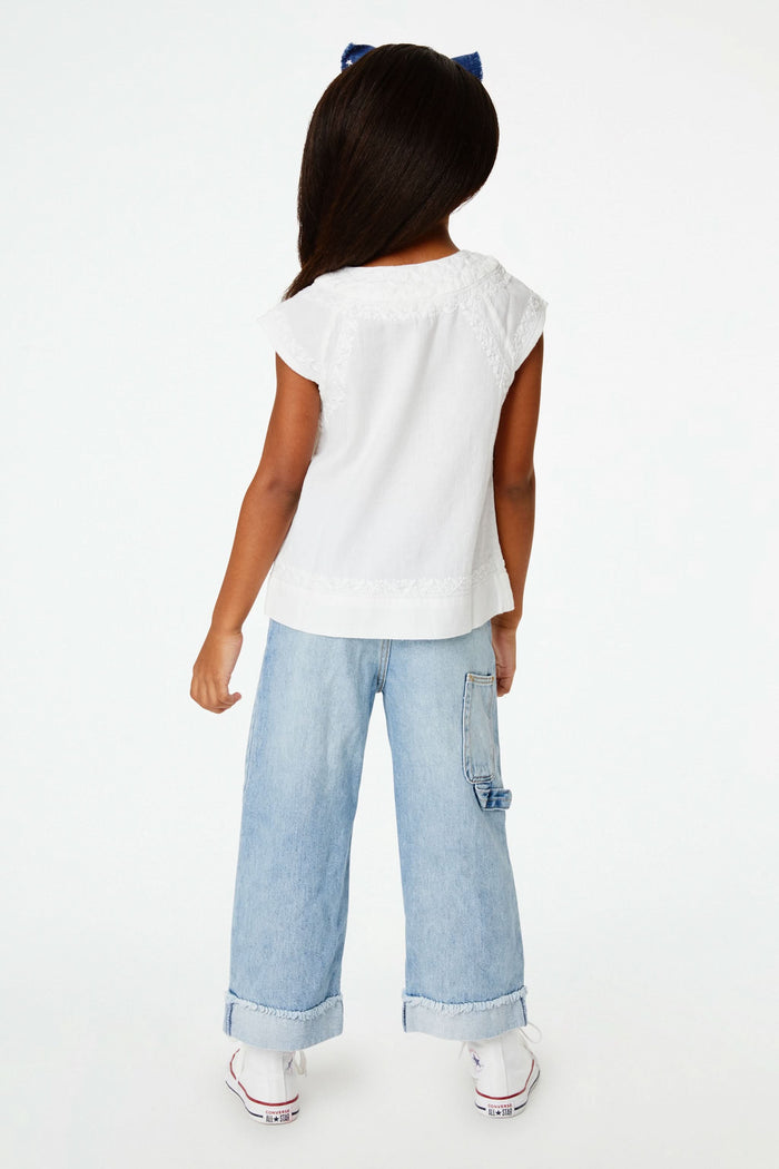 A young girl with long dark hair wears a blue headband, light blue jeans, and the Roller Rabbit Girls Faith Top in white with vibrant embroidery, standing against a plain white background and smiling slightly.