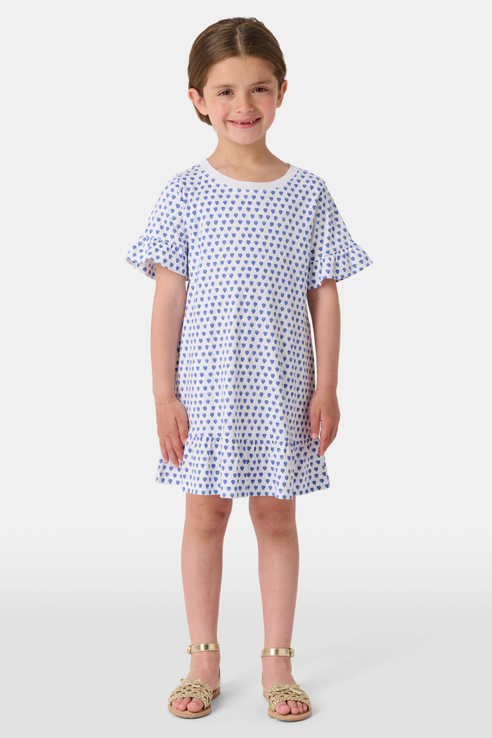 A smiling young girl faces forward in the Roller Rabbit Girls Hearts Esme Dress, featuring a white base with blue heart prints, ruffled short sleeves, and gold sandals. Her light brown hair is pulled back as she poses against a plain white background.