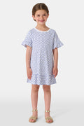 A smiling young girl faces forward in the Roller Rabbit Girls Hearts Esme Dress, featuring a white base with blue heart prints, ruffled short sleeves, and gold sandals. Her light brown hair is pulled back as she poses against a plain white background.