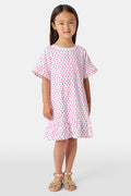 A young girl stands smiling in the Roller Rabbit Girls Hearts Esme Dress, a casual cotton T-shirt dress with pink heart patterns, short ruffled sleeves, and ruffled trim. She wears gold sandals and has long brown hair against a plain light background.
