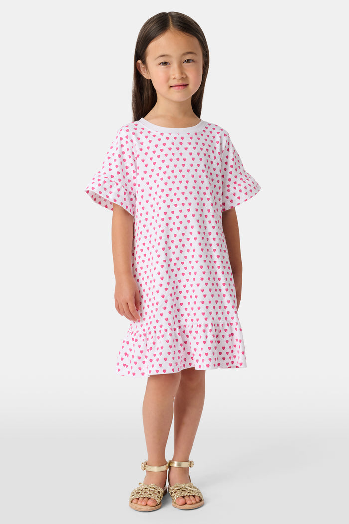 A young girl stands smiling in the Roller Rabbit Girls Hearts Esme Dress, a casual cotton T-shirt dress with pink heart patterns, short ruffled sleeves, and ruffled trim. She wears gold sandals and has long brown hair against a plain light background.