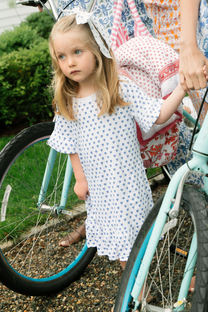 A smiling young girl faces forward in the Roller Rabbit Girls Hearts Esme Dress, featuring a white base with blue heart prints, ruffled short sleeves, and gold sandals. Her light brown hair is pulled back as she poses against a plain white background.