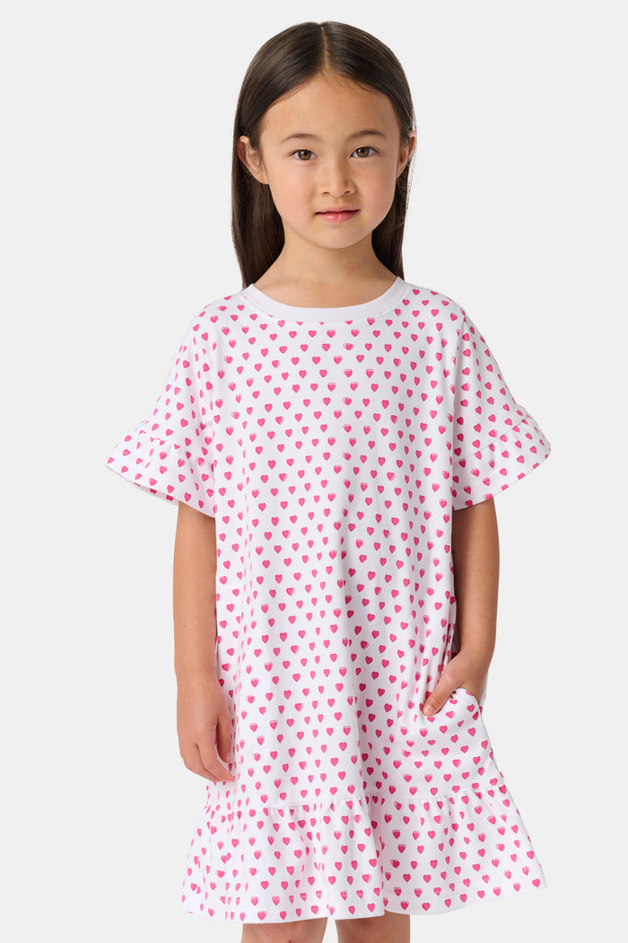 A young girl stands smiling in the Roller Rabbit Girls Hearts Esme Dress, a casual cotton T-shirt dress with pink heart patterns, short ruffled sleeves, and ruffled trim. She wears gold sandals and has long brown hair against a plain light background.