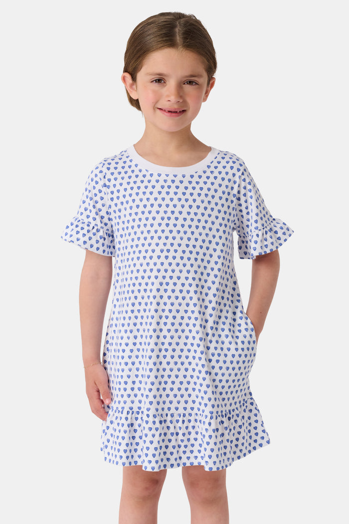 A smiling young girl faces forward in the Roller Rabbit Girls Hearts Esme Dress, featuring a white base with blue heart prints, ruffled short sleeves, and gold sandals. Her light brown hair is pulled back as she poses against a plain white background.