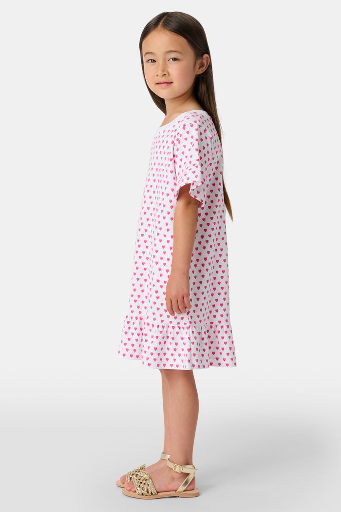 A young girl stands smiling in the Roller Rabbit Girls Hearts Esme Dress, a casual cotton T-shirt dress with pink heart patterns, short ruffled sleeves, and ruffled trim. She wears gold sandals and has long brown hair against a plain light background.