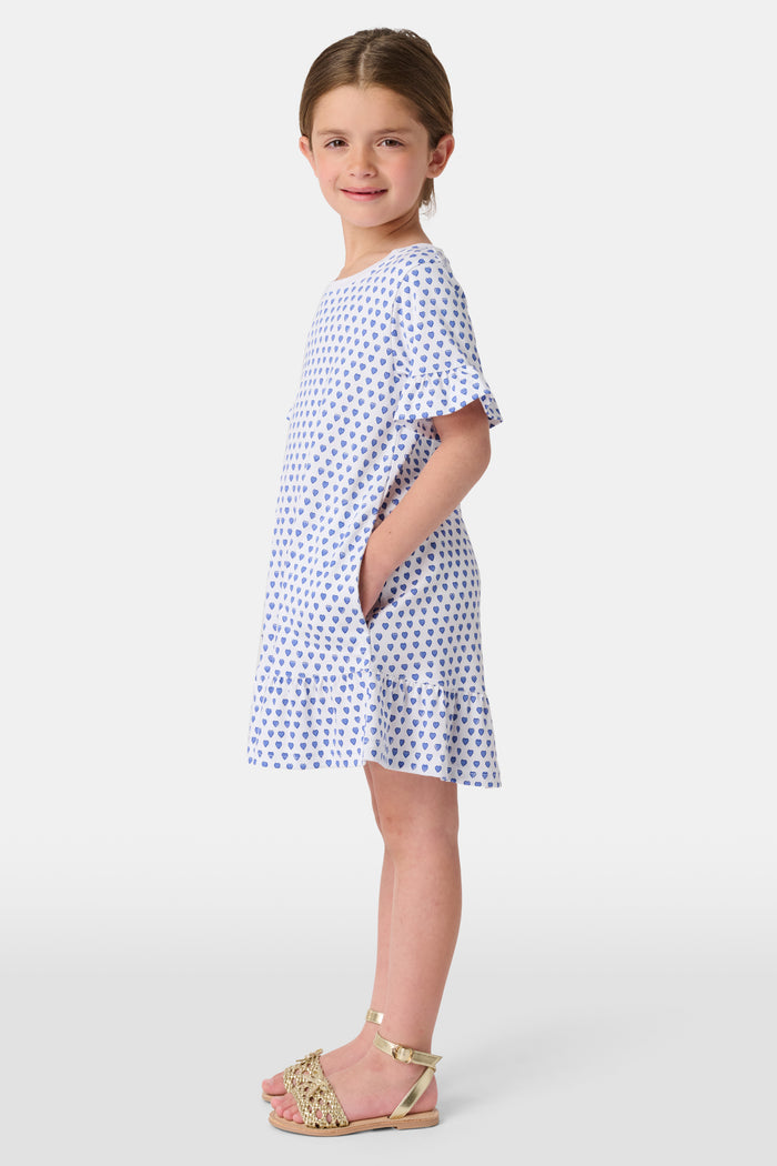A smiling young girl faces forward in the Roller Rabbit Girls Hearts Esme Dress, featuring a white base with blue heart prints, ruffled short sleeves, and gold sandals. Her light brown hair is pulled back as she poses against a plain white background.