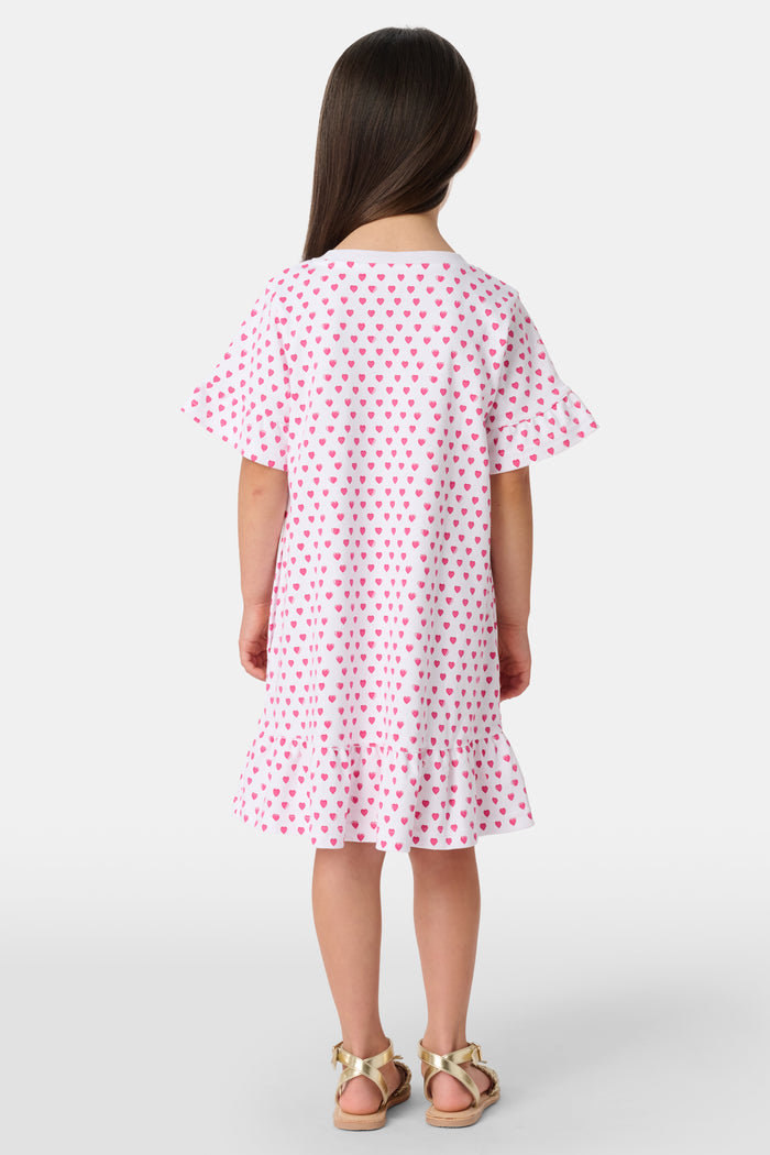 A young girl stands smiling in the Roller Rabbit Girls Hearts Esme Dress, a casual cotton T-shirt dress with pink heart patterns, short ruffled sleeves, and ruffled trim. She wears gold sandals and has long brown hair against a plain light background.