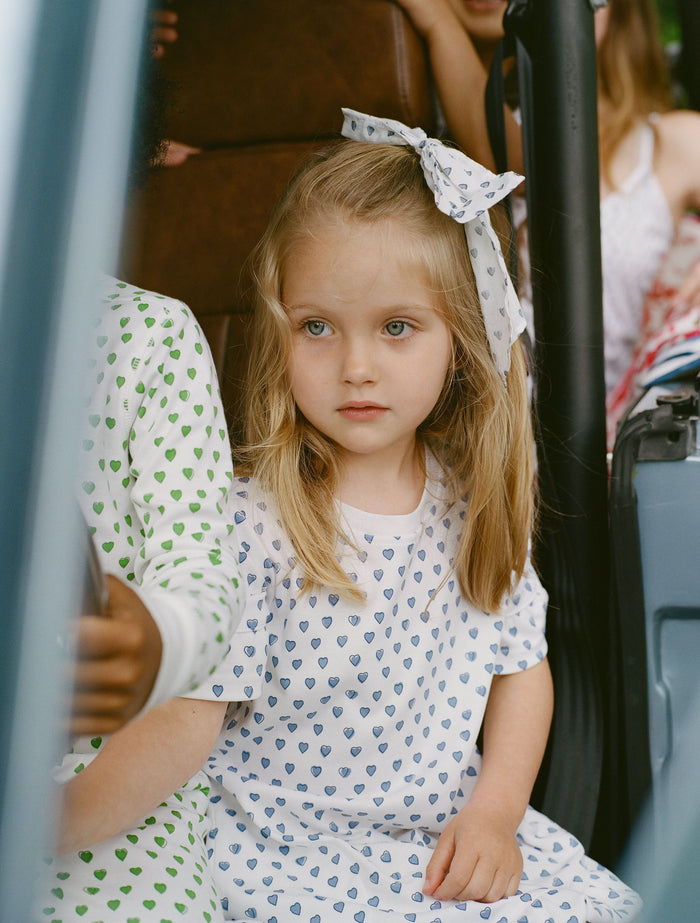 A smiling young girl faces forward in the Roller Rabbit Girls Hearts Esme Dress, featuring a white base with blue heart prints, ruffled short sleeves, and gold sandals. Her light brown hair is pulled back as she poses against a plain white background.