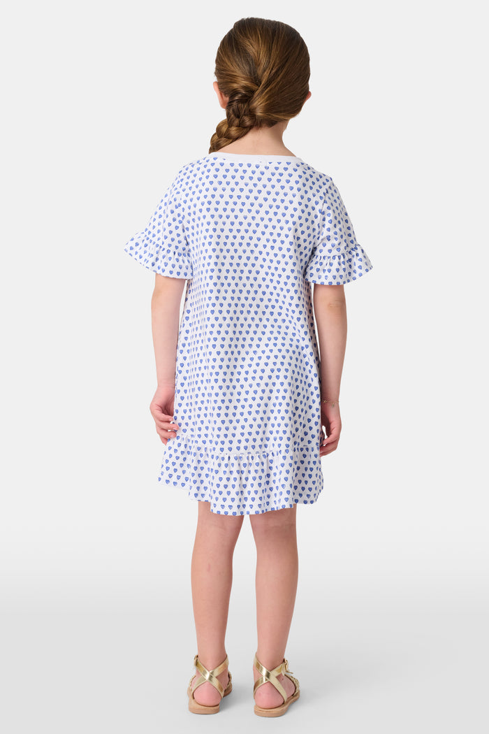 A smiling young girl faces forward in the Roller Rabbit Girls Hearts Esme Dress, featuring a white base with blue heart prints, ruffled short sleeves, and gold sandals. Her light brown hair is pulled back as she poses against a plain white background.