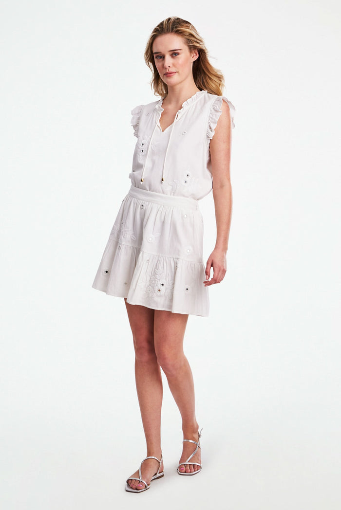 A woman models the Roller Rabbit Gloriosa Embroidery Fleet Skirt, featuring floral embroidery and eyelet details. The image captures her from waist to mid-thigh, showcasing the skirt’s delicate cotton design with one hand in her pocket.