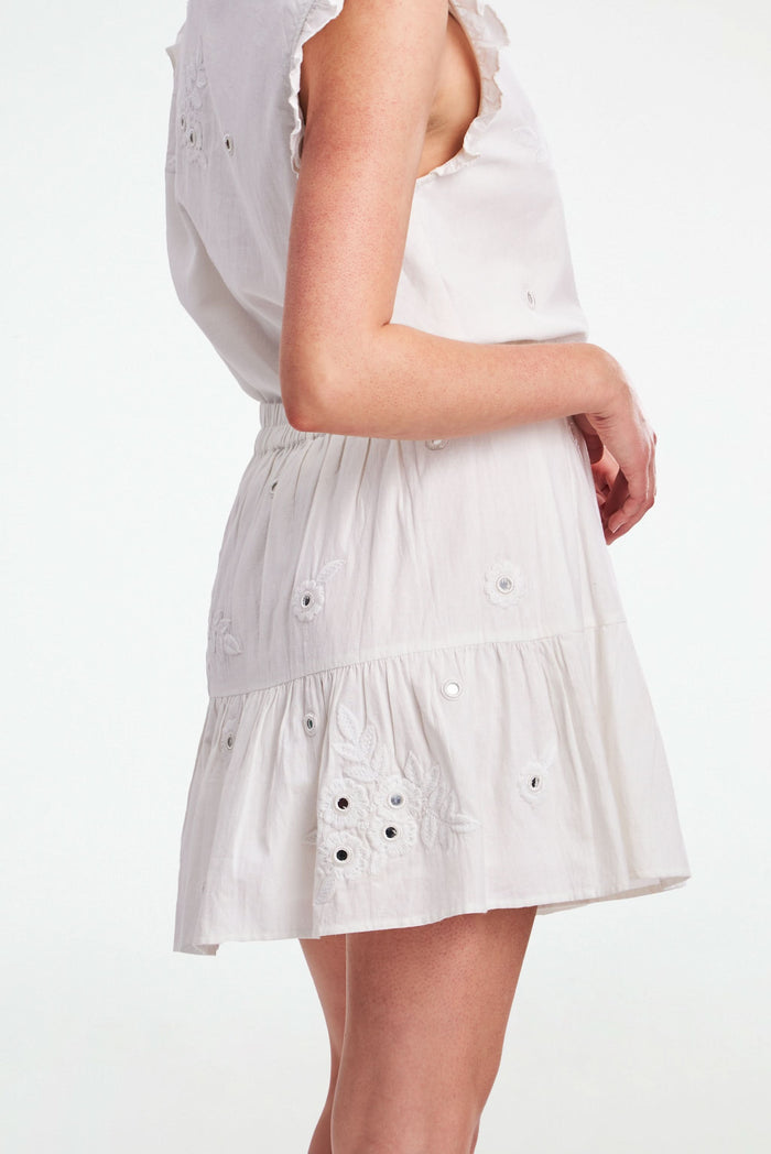 A woman models the Roller Rabbit Gloriosa Embroidery Fleet Skirt, featuring floral embroidery and eyelet details. The image captures her from waist to mid-thigh, showcasing the skirt’s delicate cotton design with one hand in her pocket.