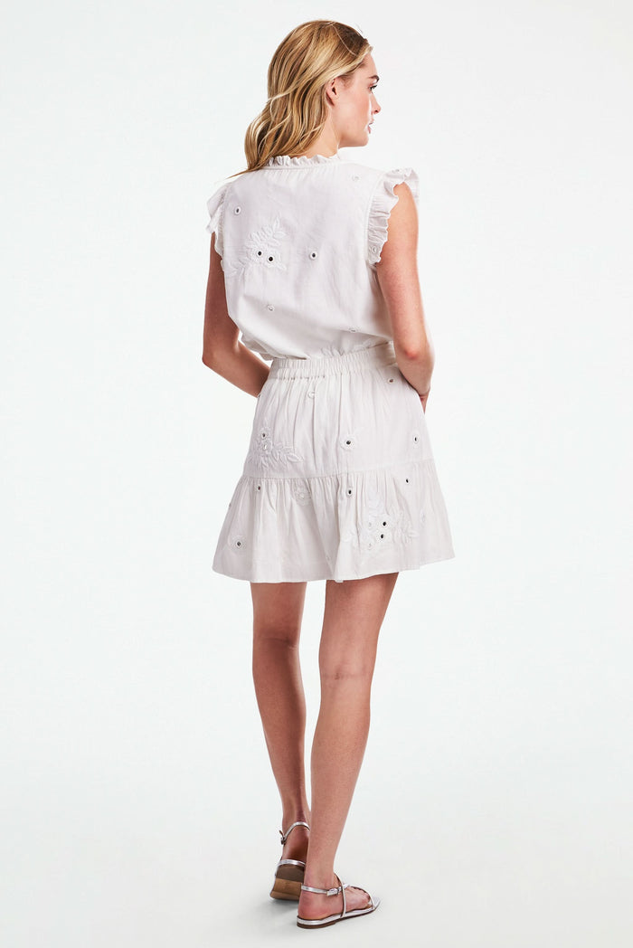 A woman models the Roller Rabbit Gloriosa Embroidery Fleet Skirt, featuring floral embroidery and eyelet details. The image captures her from waist to mid-thigh, showcasing the skirt’s delicate cotton design with one hand in her pocket.