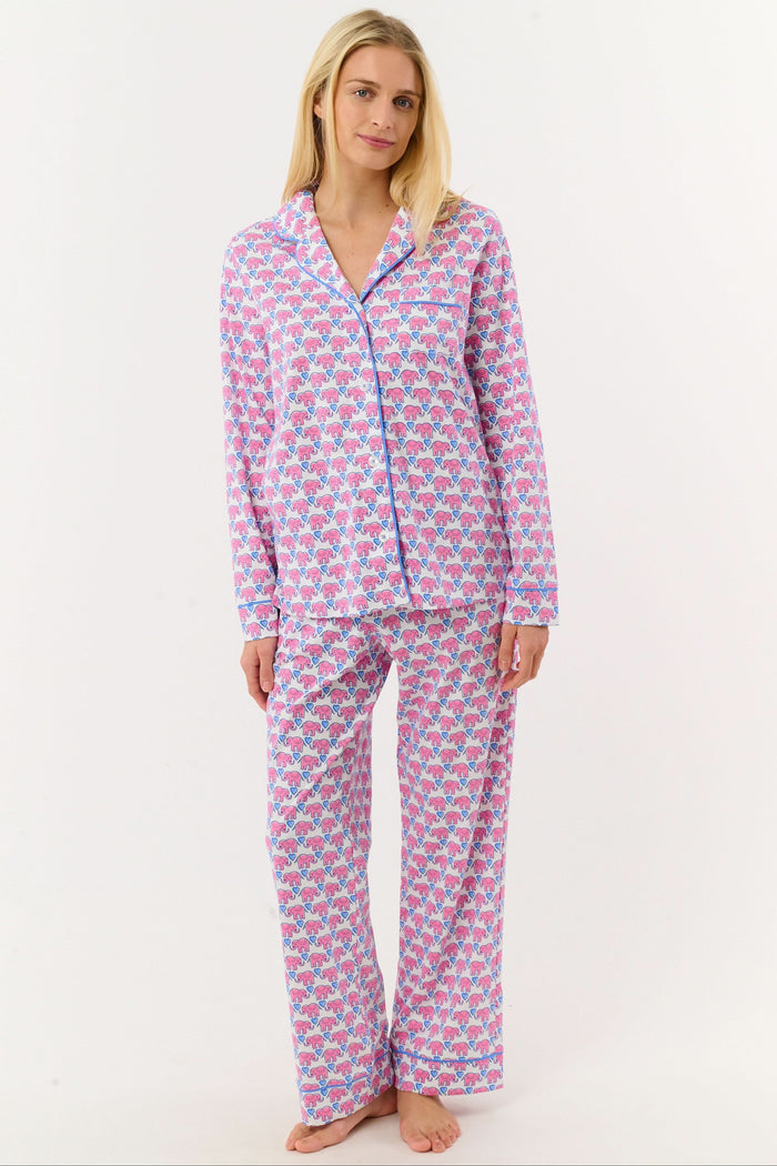 A woman stands barefoot against a white background, wearing Roller Rabbit Hathi Hearts Long Sleeve Polo Pajamas featuring a colorful pink, blue, and white floral pattern.