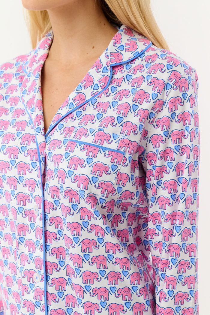 A woman stands barefoot against a white background, wearing Roller Rabbit Hathi Hearts Long Sleeve Polo Pajamas featuring a colorful pink, blue, and white floral pattern.