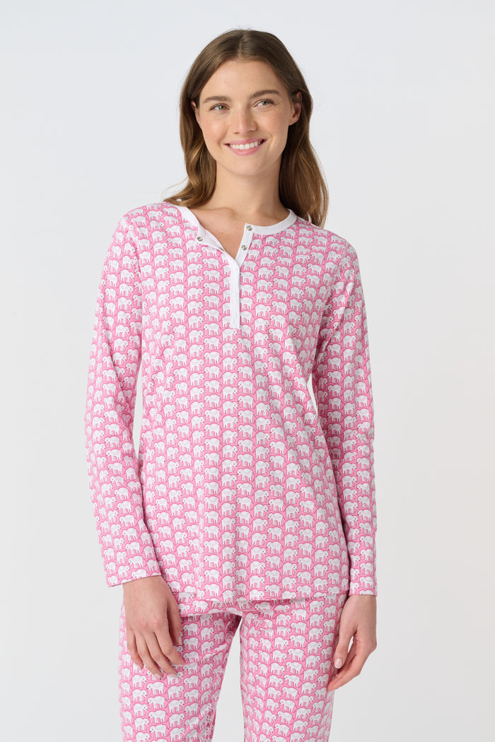 A woman stands barefoot in Roller Rabbit Hathi Pajamas—pink and white with long sleeves and pants—smiling softly against a plain white background.