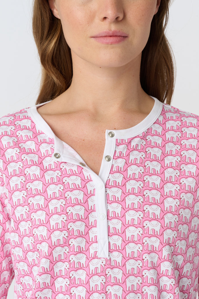 A woman stands barefoot in Roller Rabbit Hathi Pajamas—pink and white with long sleeves and pants—smiling softly against a plain white background.