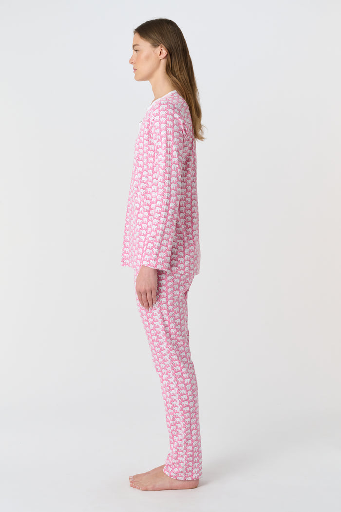 A woman stands barefoot in Roller Rabbit Hathi Pajamas—pink and white with long sleeves and pants—smiling softly against a plain white background.