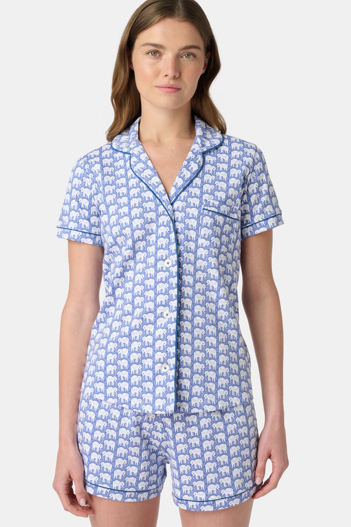 A woman wears the Hathi Polo Pajamas by Roller Rabbit, a premium cotton loungewear set with blue and white elephant patterns, including a collared button-up top and shorts, against a plain light background.