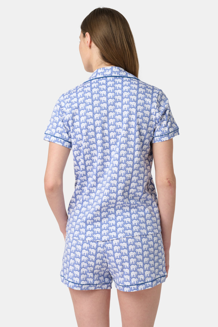 A woman wears the Hathi Polo Pajamas by Roller Rabbit, a premium cotton loungewear set with blue and white elephant patterns, including a collared button-up top and shorts, against a plain light background.