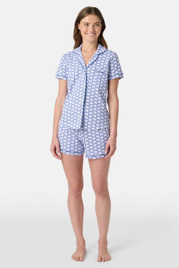 A woman wears the Hathi Polo Pajamas by Roller Rabbit, a premium cotton loungewear set with blue and white elephant patterns, including a collared button-up top and shorts, against a plain light background.