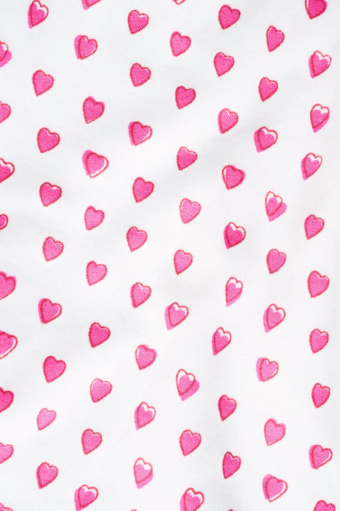 A brown poodle sits on a white background wearing Roller Rabbit's Hearts Dog Pajamas, adorable Pima Cotton pet PJs featuring small pink heart patterns for perfect matching dog pajamas.