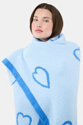 A person with straight brown hair is wrapped in the Roller Rabbit Hearts Dreams Blanket, a light blue jacquard with blue heart outlines and a bold blue stripe, standing against a plain white background.