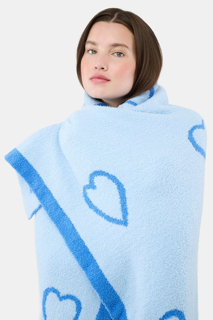 A person with straight brown hair is wrapped in the Roller Rabbit Hearts Dreams Blanket, a light blue jacquard with blue heart outlines and a bold blue stripe, standing against a plain white background.