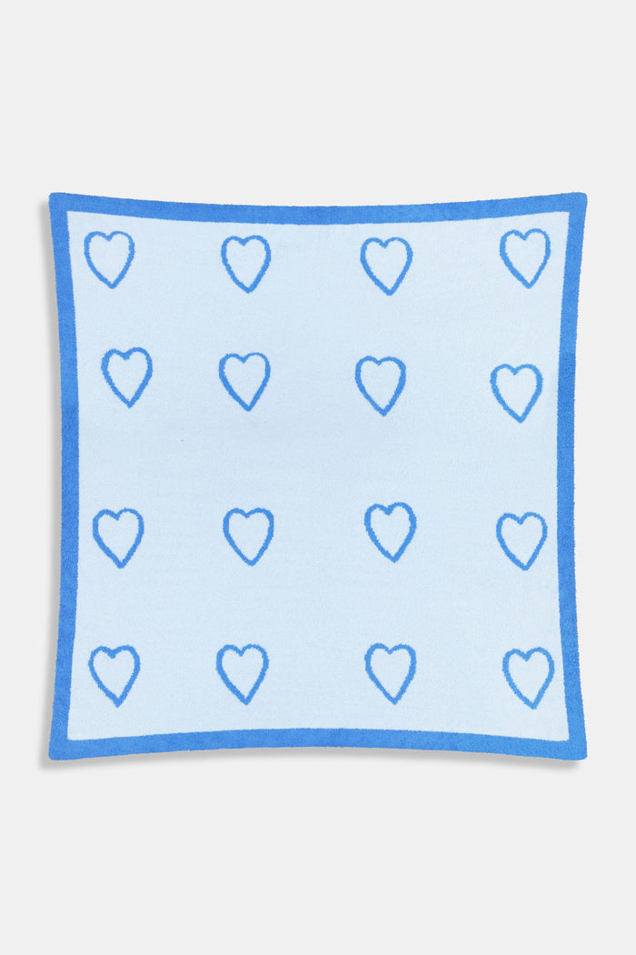 A person with straight brown hair is wrapped in the Roller Rabbit Hearts Dreams Blanket, a light blue jacquard with blue heart outlines and a bold blue stripe, standing against a plain white background.