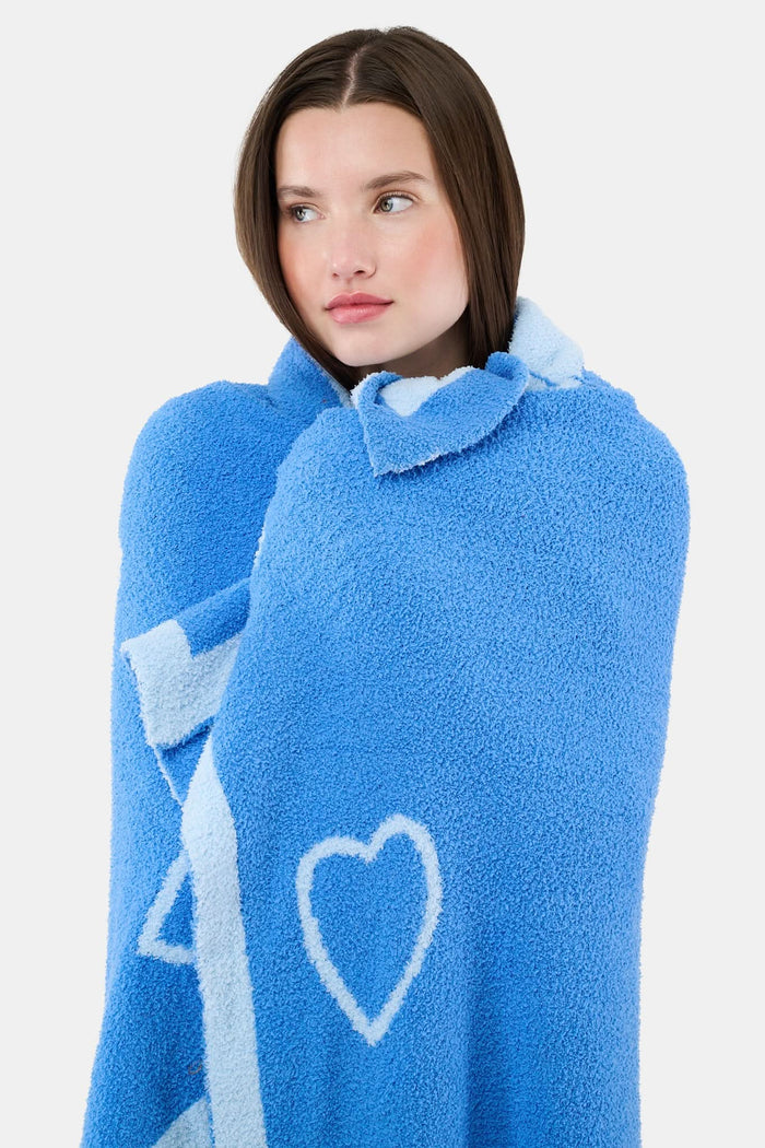 A person with straight brown hair is wrapped in the Roller Rabbit Hearts Dreams Blanket, a light blue jacquard with blue heart outlines and a bold blue stripe, standing against a plain white background.