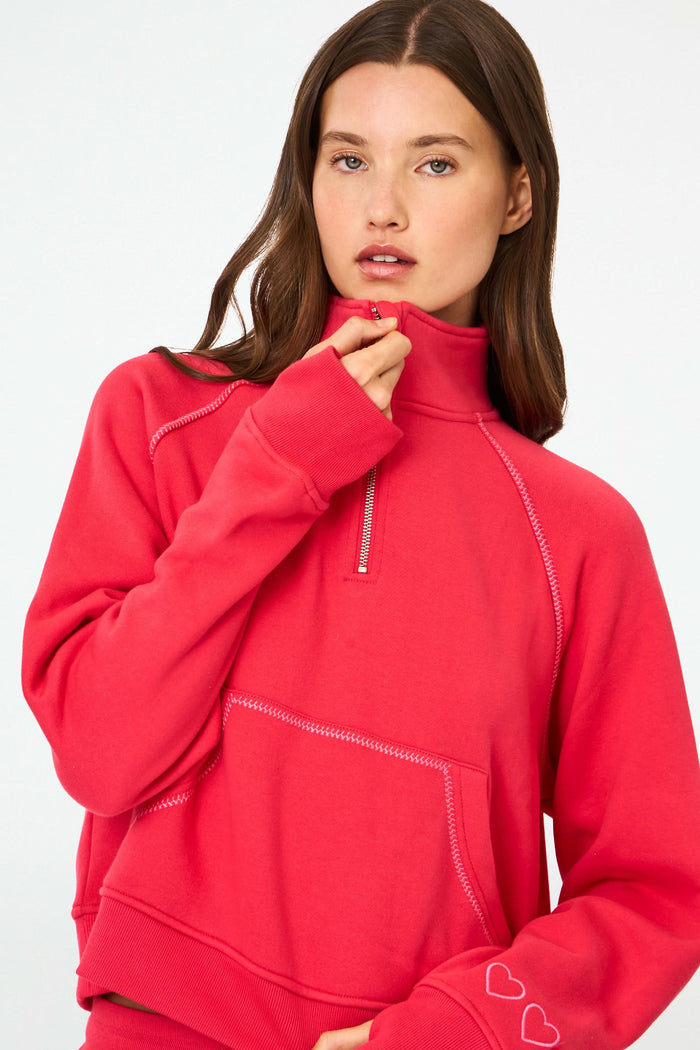 A woman with long brown hair and a neutral expression wears the cherry red Roller Rabbit Hearts Embroidered Hudson Sweatshirt, a cotton fleece half-zip pullover with a front pocket and small heart outlines on the sleeve.