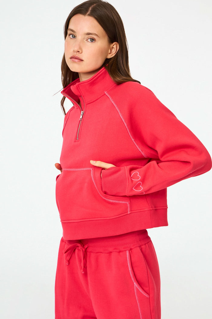 A woman with long brown hair and a neutral expression wears the cherry red Roller Rabbit Hearts Embroidered Hudson Sweatshirt, a cotton fleece half-zip pullover with a front pocket and small heart outlines on the sleeve.