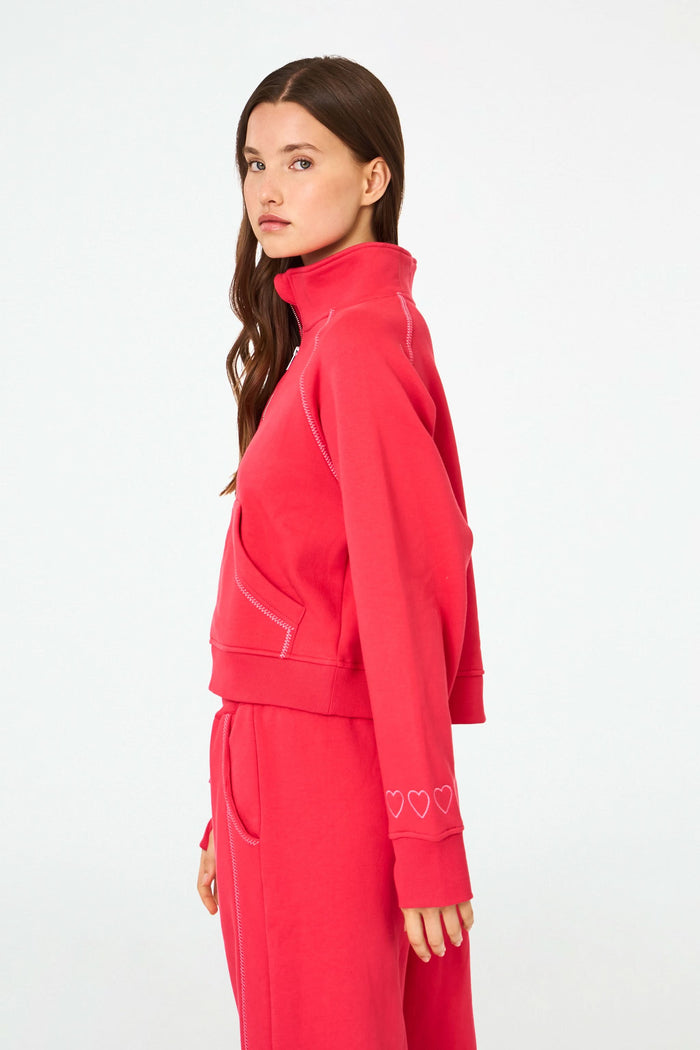 A woman with long brown hair and a neutral expression wears the cherry red Roller Rabbit Hearts Embroidered Hudson Sweatshirt, a cotton fleece half-zip pullover with a front pocket and small heart outlines on the sleeve.