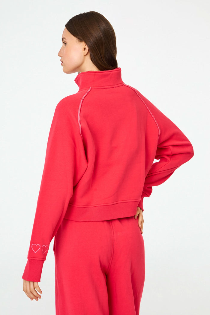 A woman with long brown hair and a neutral expression wears the cherry red Roller Rabbit Hearts Embroidered Hudson Sweatshirt, a cotton fleece half-zip pullover with a front pocket and small heart outlines on the sleeve.
