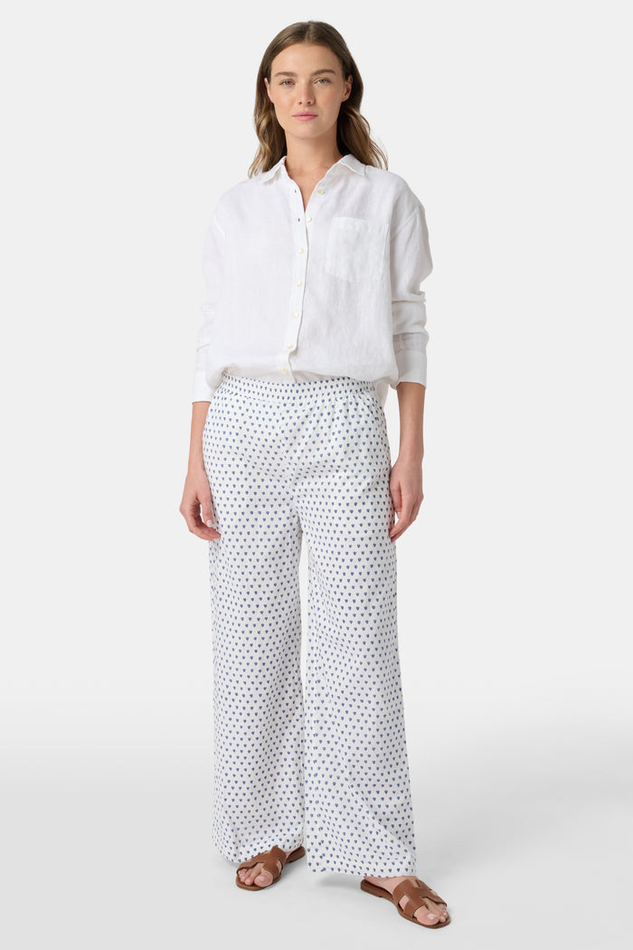 A person stands facing forward, wearing Roller Rabbit’s Hearts Kaylee Pant and a white cotton button-up shirt with brown flat sandals against a plain light background.