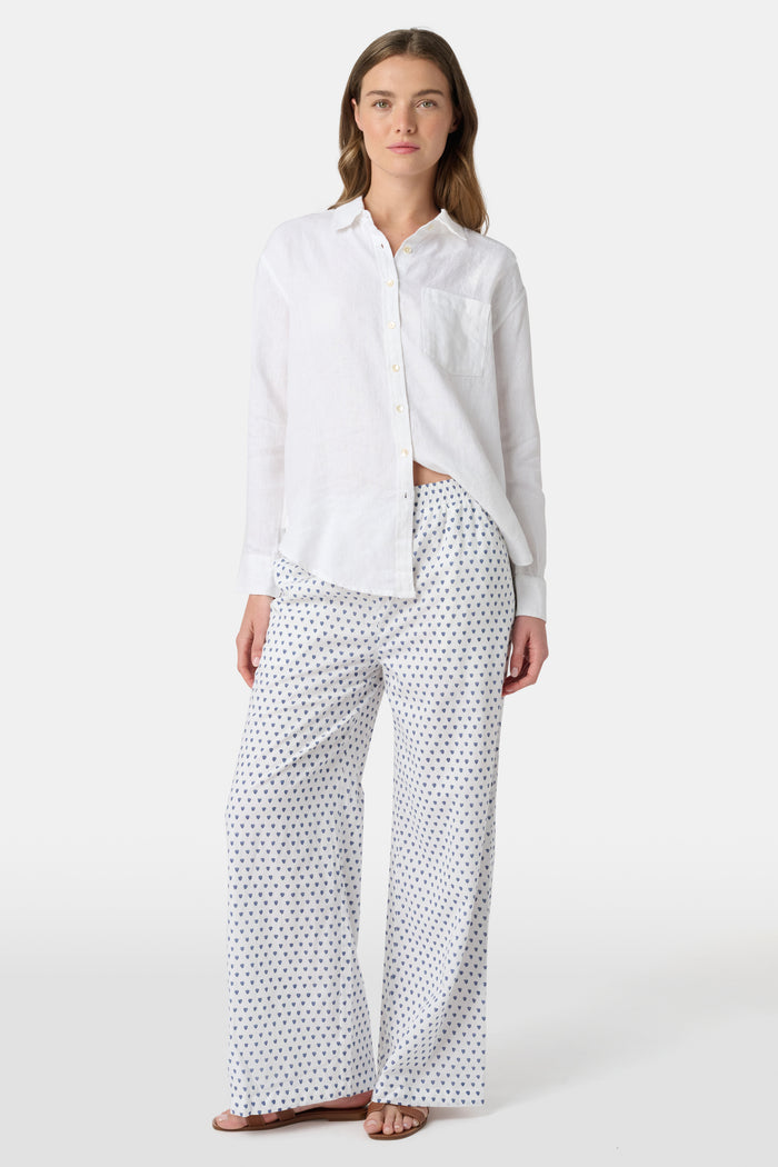 A person stands facing forward, wearing Roller Rabbit’s Hearts Kaylee Pant and a white cotton button-up shirt with brown flat sandals against a plain light background.