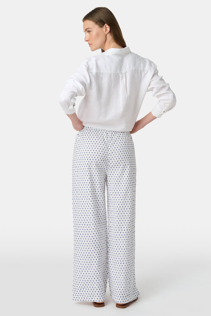 A person stands facing forward, wearing Roller Rabbit’s Hearts Kaylee Pant and a white cotton button-up shirt with brown flat sandals against a plain light background.