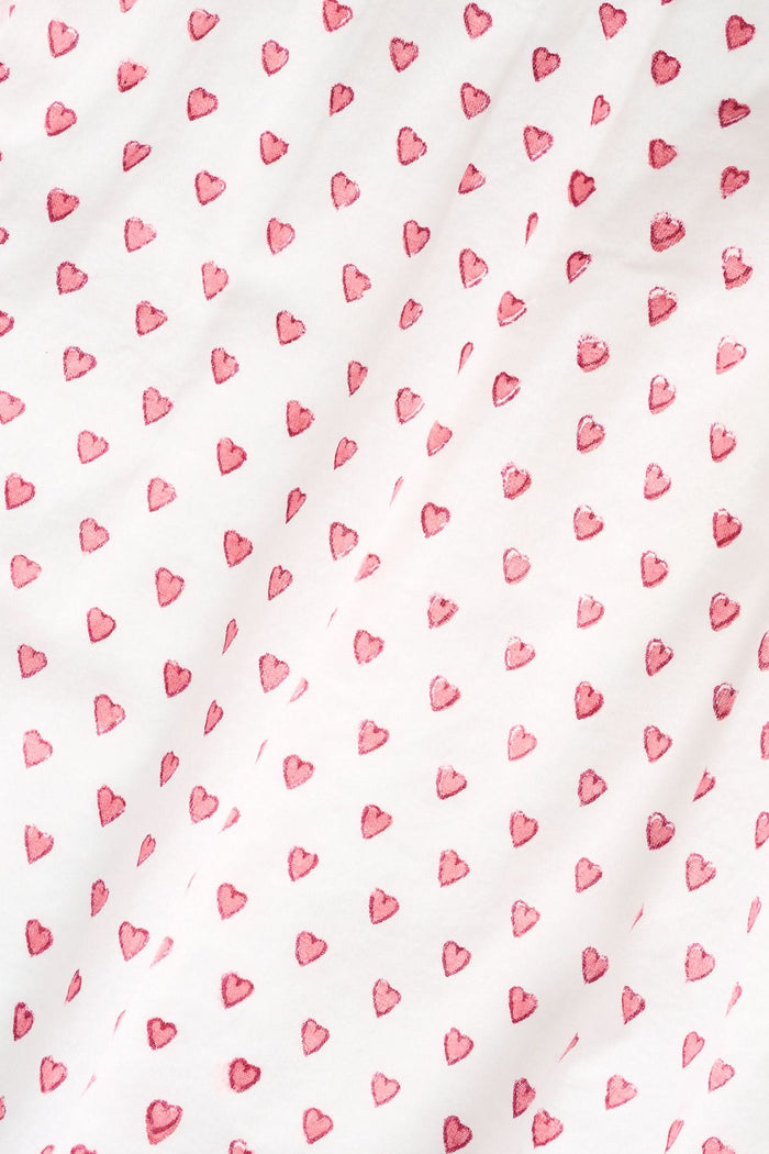 A person stands barefoot in the Roller Rabbit Hearts Christy Set—a light pink lounge set with small red hearts, featuring a belted wrap top and loose-fitting pants—against a plain white background.
