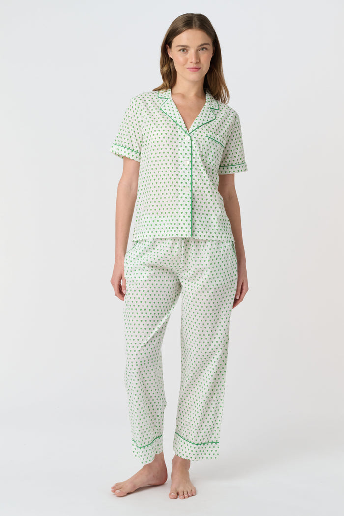 A woman stands barefoot against a plain background, wearing the Roller Rabbit Hearts Liza Set—a matching pajama set in soft 60's Cambric Cotton, featuring short sleeves, long pants, a white base with green hearts, and green piping edges.
