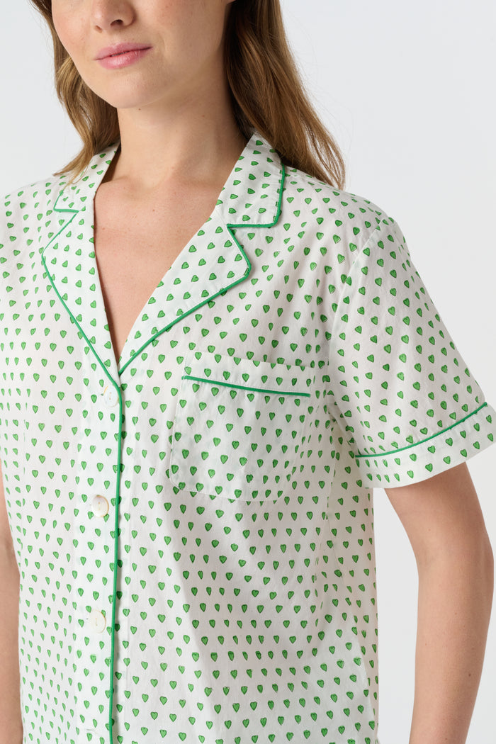 A woman stands barefoot against a plain background, wearing the Roller Rabbit Hearts Liza Set—a matching pajama set in soft 60's Cambric Cotton, featuring short sleeves, long pants, a white base with green hearts, and green piping edges.