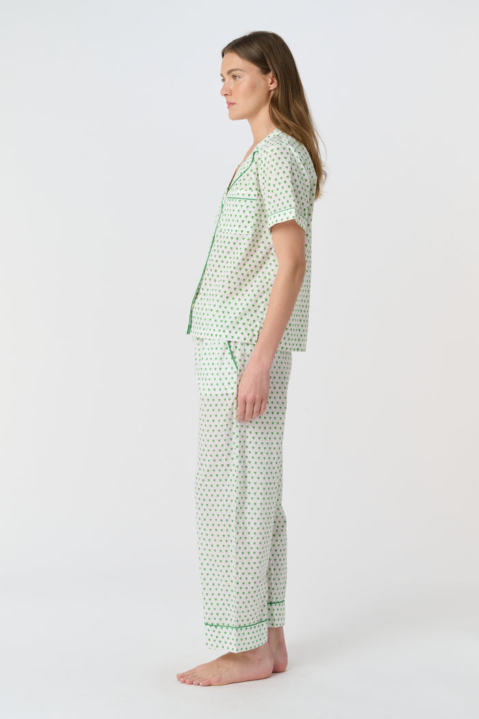 A woman stands barefoot against a plain background, wearing the Roller Rabbit Hearts Liza Set—a matching pajama set in soft 60's Cambric Cotton, featuring short sleeves, long pants, a white base with green hearts, and green piping edges.