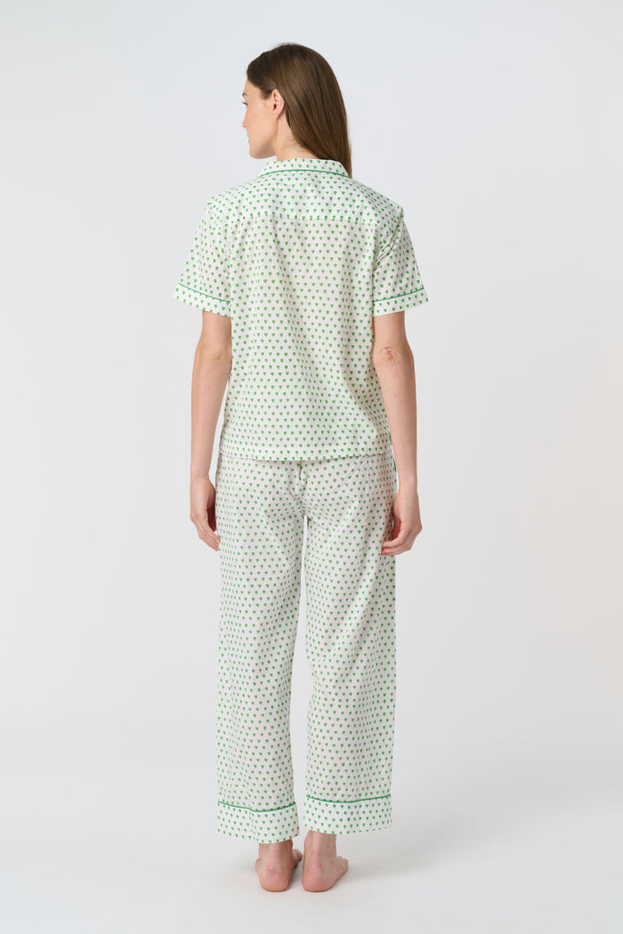 A woman stands barefoot against a plain background, wearing the Roller Rabbit Hearts Liza Set—a matching pajama set in soft 60's Cambric Cotton, featuring short sleeves, long pants, a white base with green hearts, and green piping edges.
