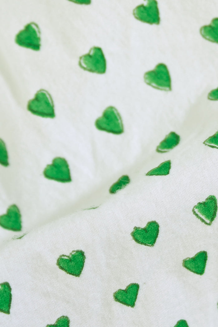 A woman stands barefoot against a plain background, wearing the Roller Rabbit Hearts Liza Set—a matching pajama set in soft 60's Cambric Cotton, featuring short sleeves, long pants, a white base with green hearts, and green piping edges.