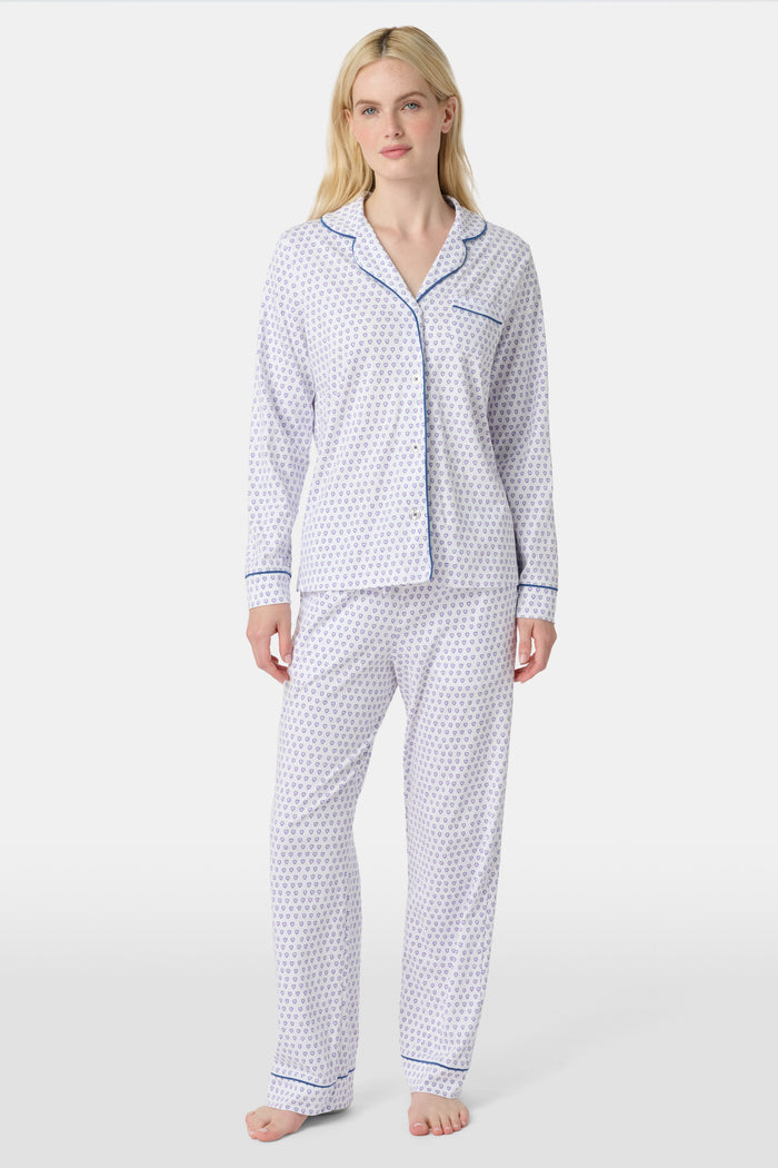 A woman stands barefoot in Roller Rabbit’s Hearts Long Sleeve Polo Pajamas, a Pima cotton set featuring a button-up top and long pants with blue piping and small blue heart patterns on a white background.