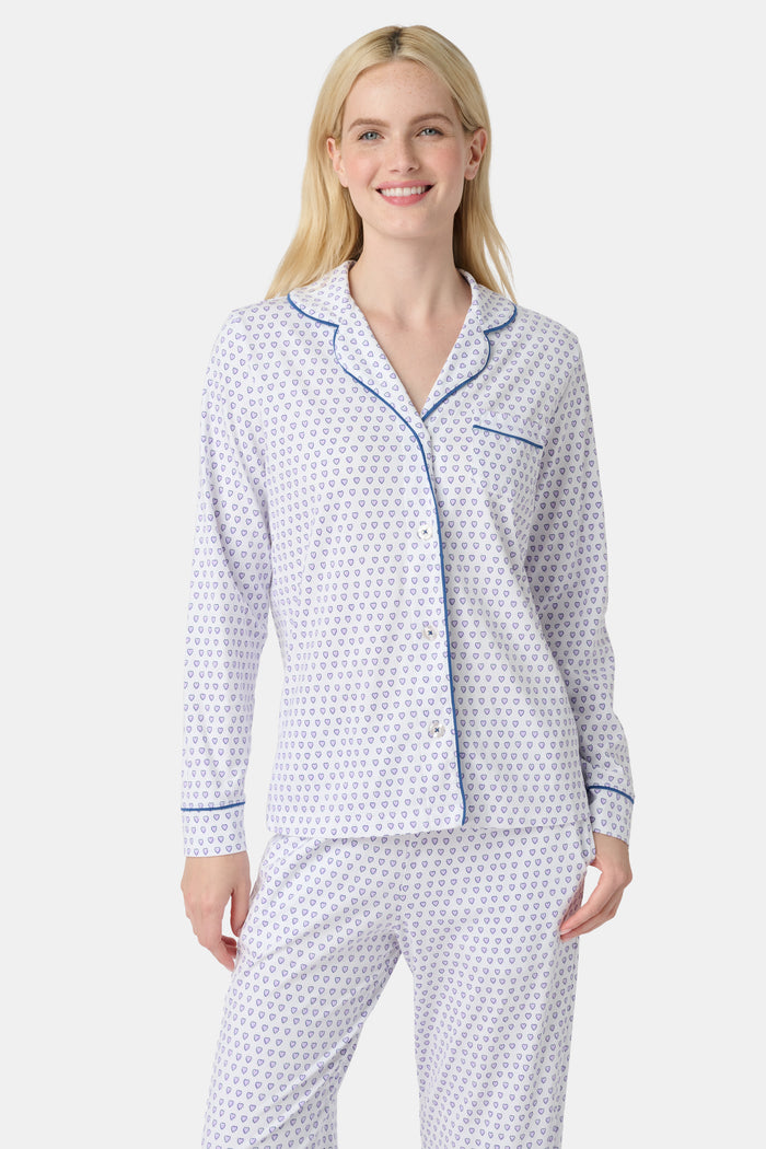 A woman stands barefoot in Roller Rabbit’s Hearts Long Sleeve Polo Pajamas, a Pima cotton set featuring a button-up top and long pants with blue piping and small blue heart patterns on a white background.