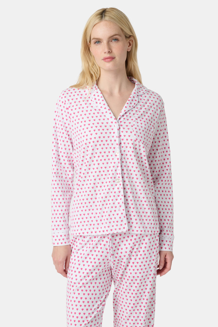A woman stands barefoot in Roller Rabbit's Hearts Long Sleeve Polo Pajamas, a light pink Pima cotton set with a collared button-up top and matching pants, both decorated with small pink and purple hearts.