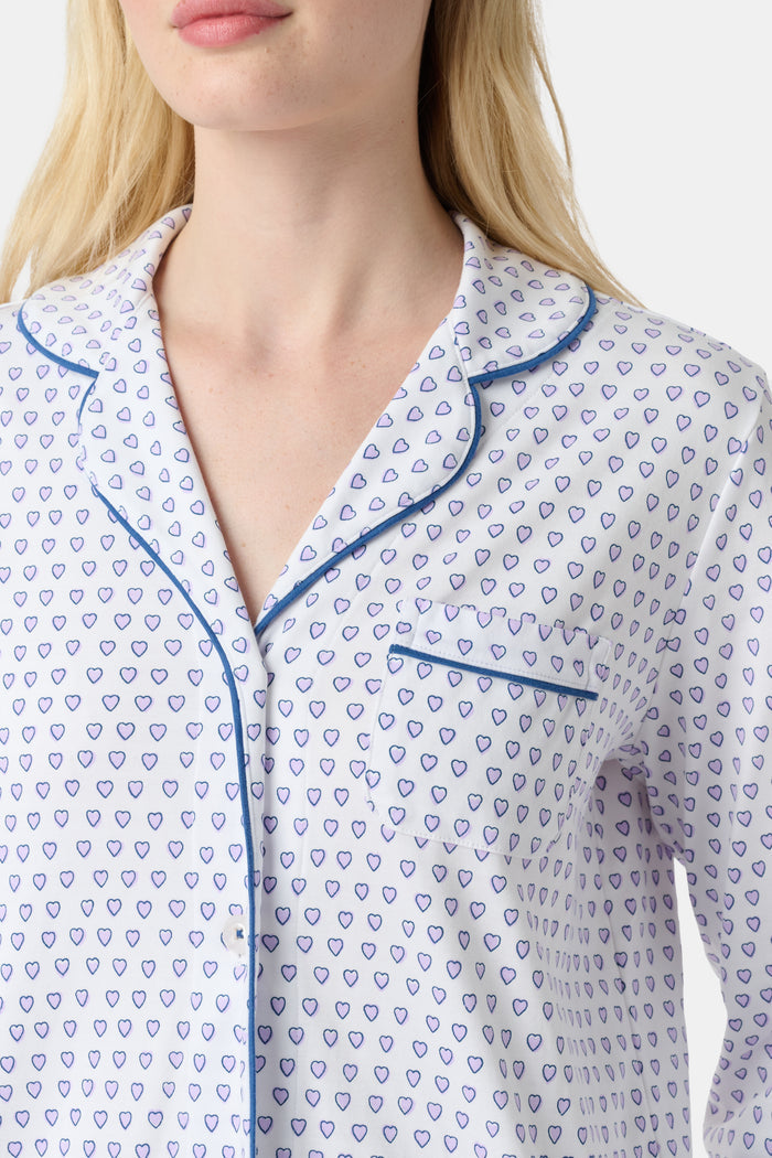 A woman stands barefoot in Roller Rabbit’s Hearts Long Sleeve Polo Pajamas, a Pima cotton set featuring a button-up top and long pants with blue piping and small blue heart patterns on a white background.
