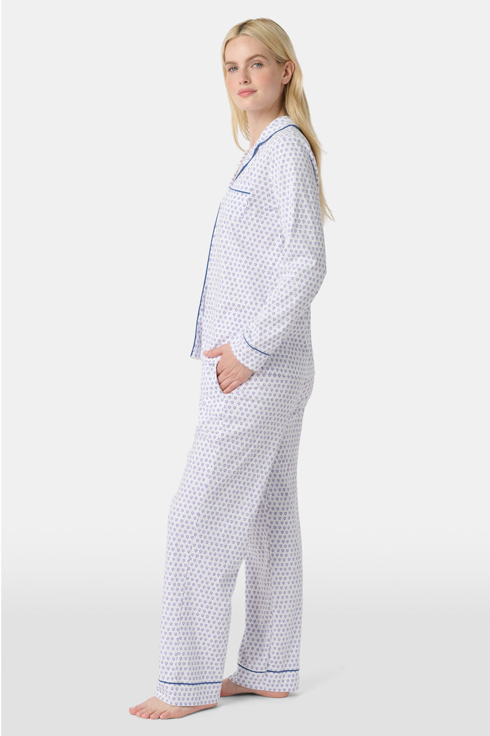 A woman stands barefoot in Roller Rabbit’s Hearts Long Sleeve Polo Pajamas, a Pima cotton set featuring a button-up top and long pants with blue piping and small blue heart patterns on a white background.